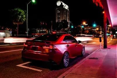 Rear three-quarter view of a dark red sport sedan wrapped in Satin Vampire Red, parked on a city street at night