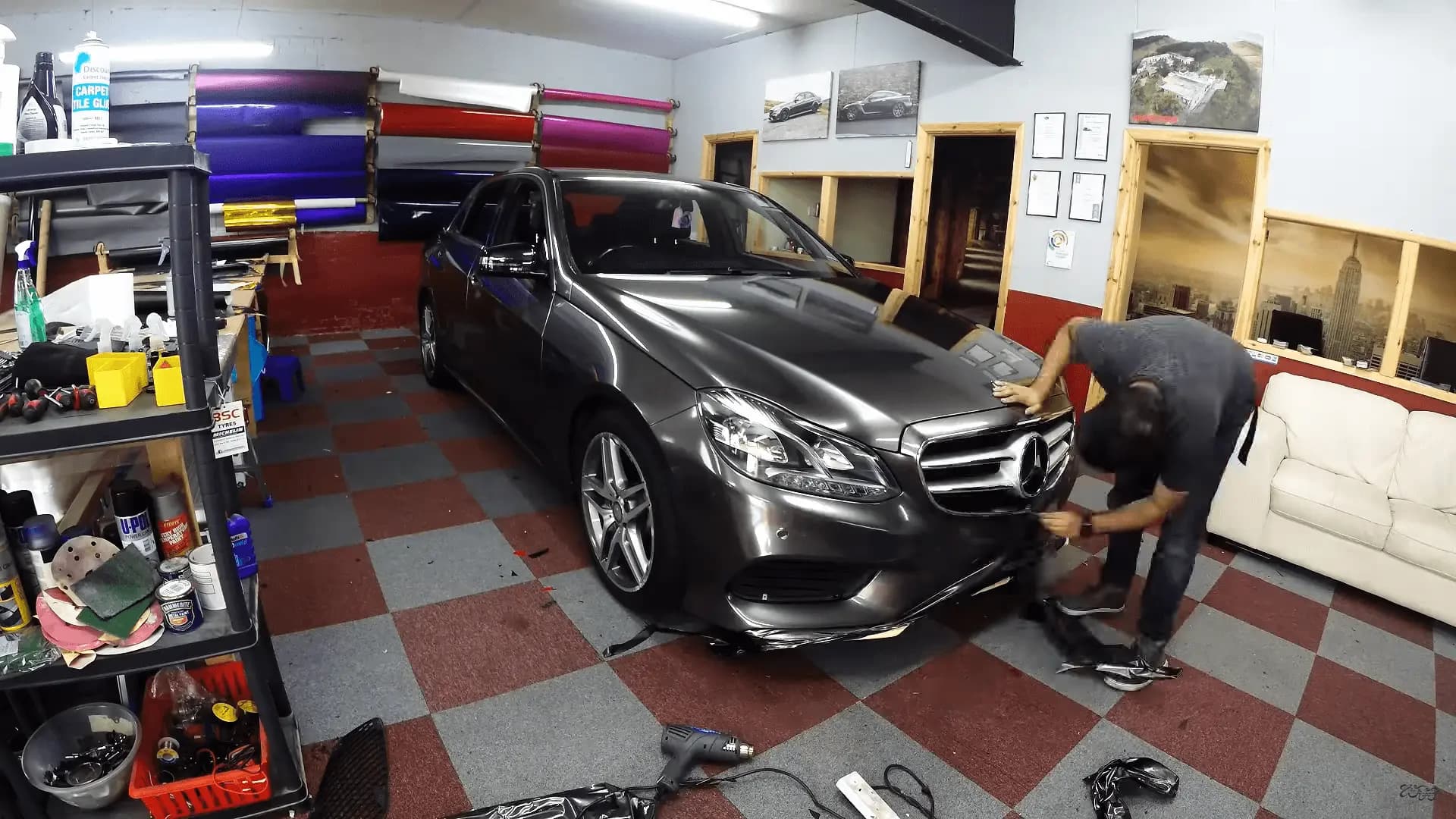 Mercedes-Benz E-Class sedan in dark metallic grey Gloss Anthracite, an installer smoothing film along the front bumper in a wrap shop