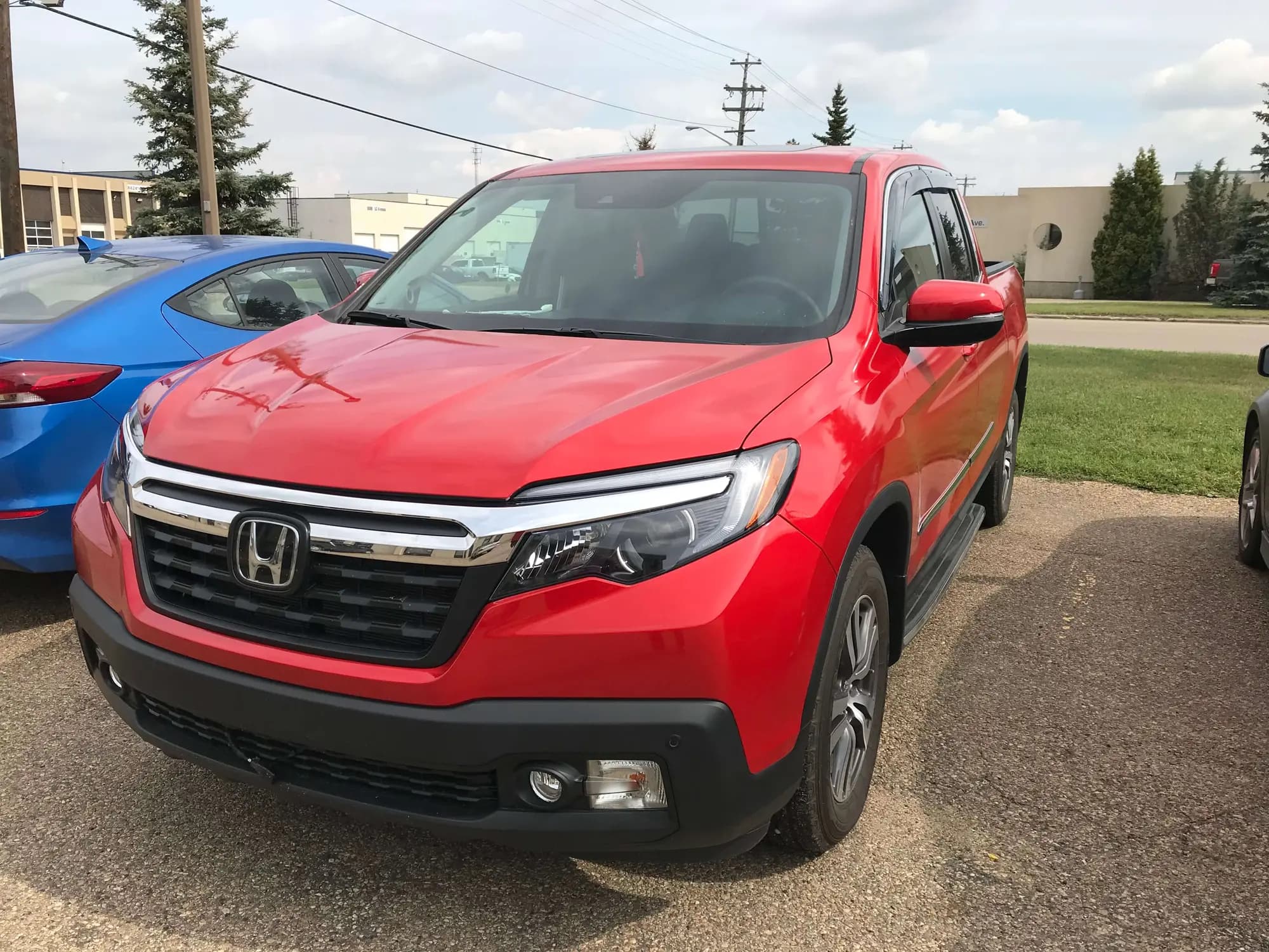 Red Honda Ridgeline crew cab pickup on a gravel lot, front three-quarter view, its gloss red hood reflecting the trees and overcast sky, shown for Gloss Dragon Fire Red