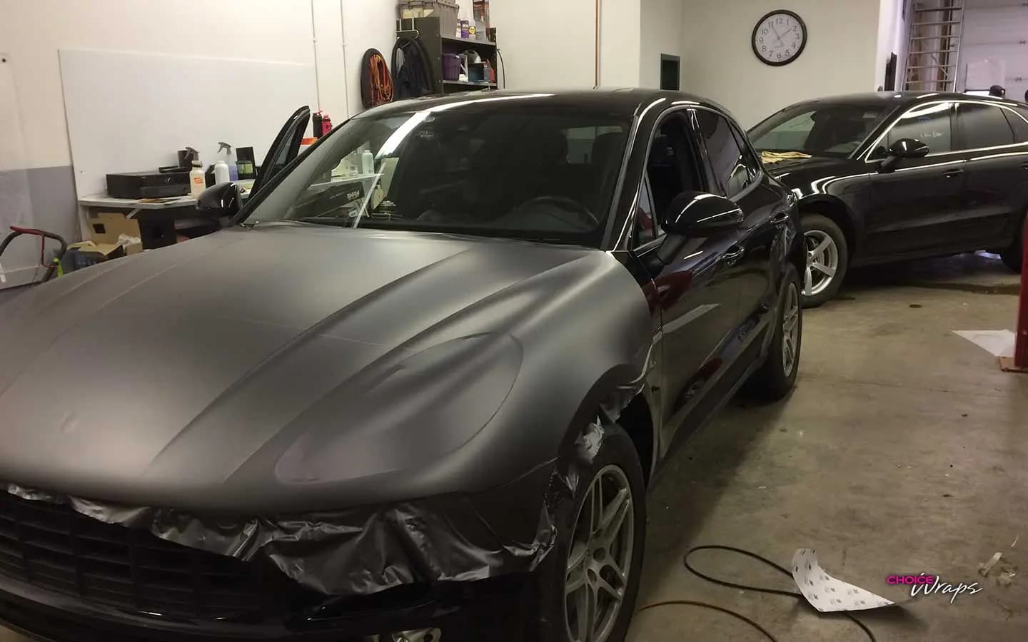 Porsche Macan in the shop bay partway through a Matte Dark Gray wrap, hood and front fender covered while the rest of the body is still black