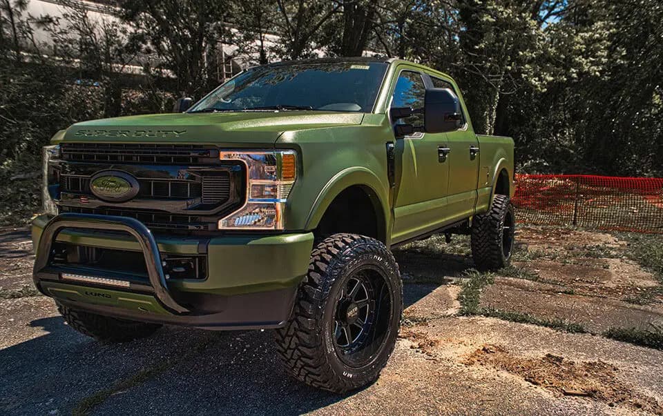 Lifted Ford Super Duty crew cab pickup wrapped in Rugged Marsh Green, parked on a gravel lot in front of trees